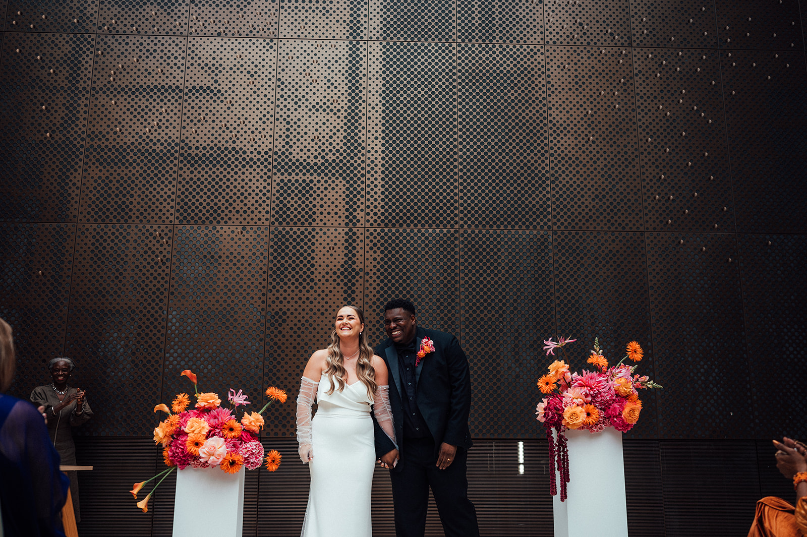 Image of a happy newlywed couple standing in between plinths that are covered in flowers in the Atrium at Hackney Town Hall